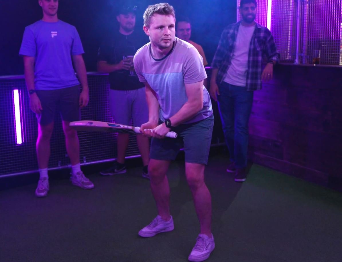 A batter plays a shot in a neon-lit cricket net while friends watch