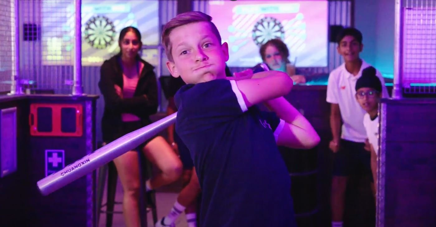 A young batter swings while friends cheer from the side of the net